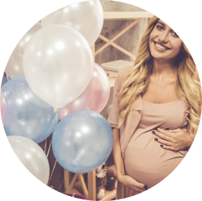 Smiling woman at a celebration holding her pregnant belly, surrounded by blue and white balloons.