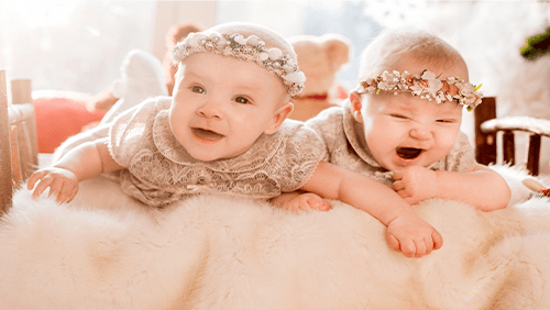 Two babies in floral headbands lying on a furry surface, one smiling and one crying.