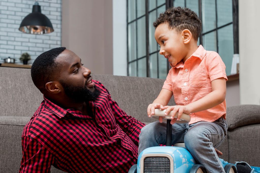 A young child sitting on a toy car converses with an attentive adult male on a sofa.