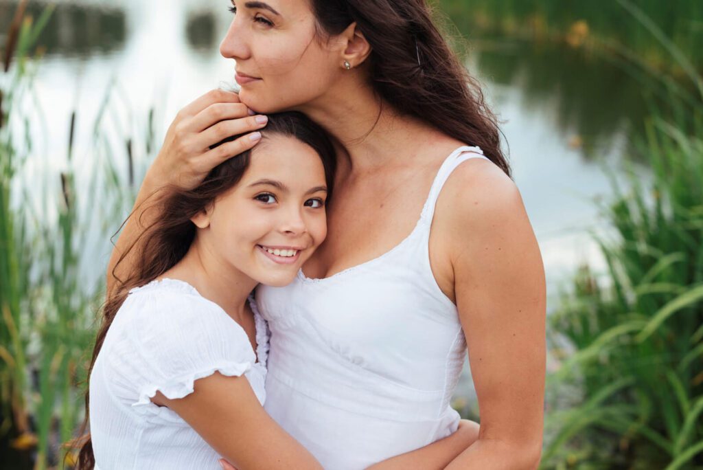 A young girl embraces her mother from behind, both smiling and looking into the distance, near a body of water.