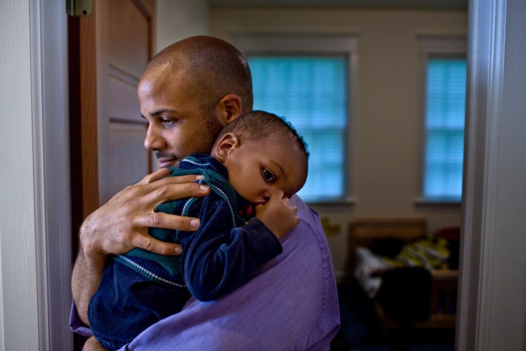 A bald man comforting a sleepy toddler in his arms inside a home.